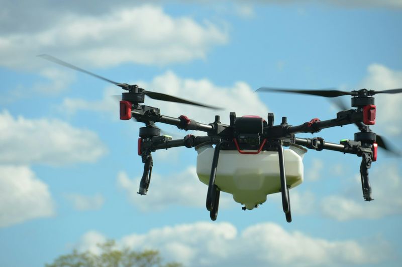 Agricultural spray drone with a white tank flying under a bright blue sky with clouds
